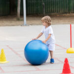 little boy rolling a big blue ball on a playground surface