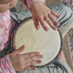 a child with a pink shirt plays a drum outside on a bench