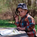 Boy wearing hat looks through magnifying glass at nature book outside