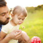 father and toddler daughter with blonde pigtails blowing out a dandelion in a park