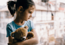 Little girl holding a teddy bear looking out a window