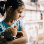 Little girl holding a teddy bear looking out a window