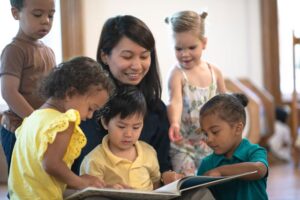 teacher smiling and reading a book to several toddlers