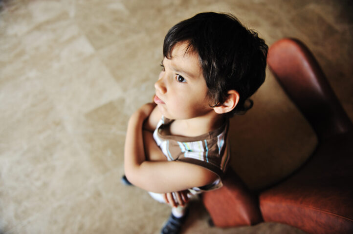 angry boy with brown hair and arms crossed standing near a chair