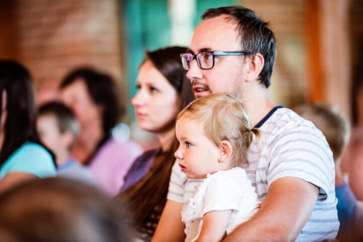 Blonde toddler girl sitting with father who is wearing glasses, near mother at outdoor event.
