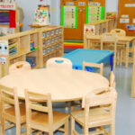 preschool classroom with wooden group table with chairs, activity centers, and shelving with learning materials