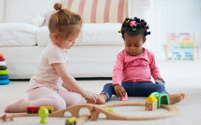 two toddler girls playing with wooden train set on floor