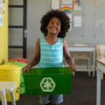 Preschool boy in blue shirt smiling holds a green recycling bin in a classroom