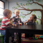 preschool teacher and two children in classroom library playing with pretend food at large wooden table with tree mural on wall in the background