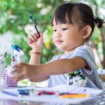 preschool girl with paintbrush at table outdoors painting.